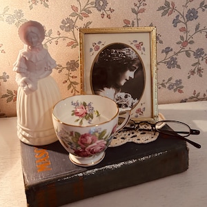 May include: A vintage porcelain figurine of a woman in a white dress, a teacup with floral pattern, a framed photo, and a pair of eyeglasses on a book with the word "MASSA" on the cover.