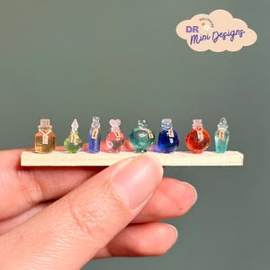 May include: Miniature glass potion bottles in various colors, including amber, green, blue, and red, are arranged on a small wooden shelf. Each bottle has a unique shape and is labeled with a tiny tag. The image is labeled with the text "DR Mini Designs".