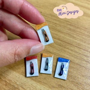 May include: Four miniature utility knives in individual clear plastic blister packs with cardboard backings. The knives come in orange, red, silver, and blue. Each backing features the text DR Mini Designs at the top of the packaging.