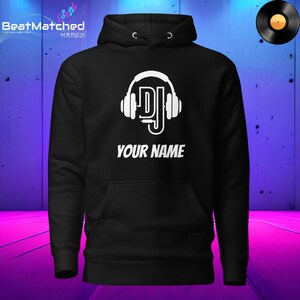 May include: Black hoodie with a white DJ graphic and the text "YOUR NAME". The design includes headphones and the letters "DJ". A vinyl record is in the upper right corner. The hoodie has a front pocket and a drawstring hood.