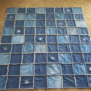 Peut inclure: Une courtepointe en patchwork de denim avec un motif quadrillé de carrés de denim bleu de différentes nuances. Certains carrés ont des poches. Des broderies de flocons de neige blancs ornent la courtepointe. La courtepointe est présentée sur un tapis de couleur neutre.