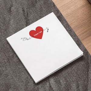 May include: A stack of white notepaper with a red heart graphic and the words "Nurses Care Always" in cursive. A syringe and other small graphics are also present. The notepaper is on a gray surface.