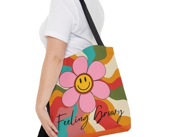 May include: A tote bag with a retro-style design featuring a pink flower with a yellow smiley face. The bag has a black strap and the text "Feeling Groovy" in a black script font.
