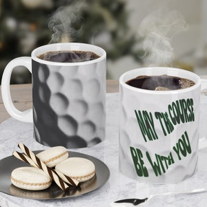 Puede incluir: Dos tazas de café blancas con asas negras. Una taza tiene un diseño de bola de golf y la otra taza tiene el texto "Que el campo esté contigo" en letras verdes.