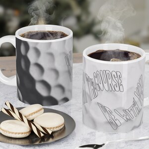 May include: Two white ceramic coffee mugs with black coffee. One mug has a golf ball texture and the text "BE THE COURSE BE WITH YOU". The other mug has a golf ball texture.