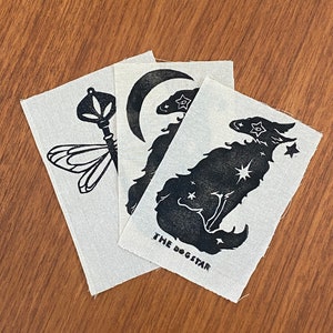 Pode incluir: Três patches de tecido com desenhos de tinta preta. O primeiro patch apresenta uma libélula com uma chave. O segundo patch apresenta uma lua crescente e uma estrela. O terceiro patch apresenta um lobo estilizado com estrelas e o texto "THE DOGSTAR".
