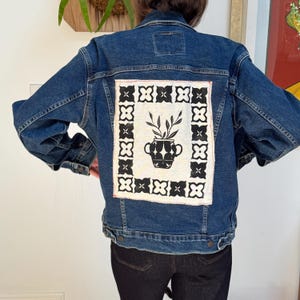 Puede incluir: Chaqueta vaquera azul oscuro con un parche decorativo en la espalda. El parche presenta un diseño en blanco y negro de un jarrón con plantas, enmarcado por un borde negro. La chaqueta tiene un diseño clásico con botones.