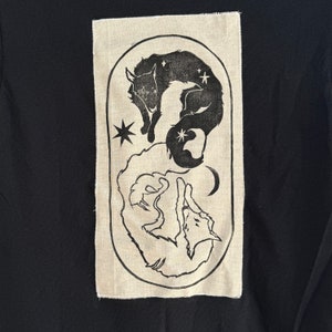 Peut inclure: Un patch en tissu beige avec de l'encre noire représentant deux animaux stylisés, un noir et un blanc, enroulés en cercle. Les animaux sont entourés d'étoiles et d'un croissant de lune.