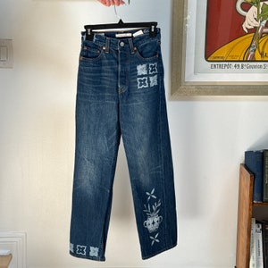 May include: Blue denim jeans with white embroidered floral designs and a potted plant motif. The jeans have a high waist and a wide leg.