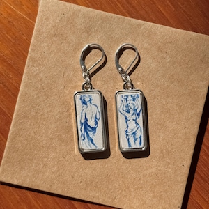 May include: A pair of silver earrings. Each earring has a rectangular pendant with a blue and white illustration of a classical figure. The figures are hand-painted with fine detail. The earrings have a lever-back closure.