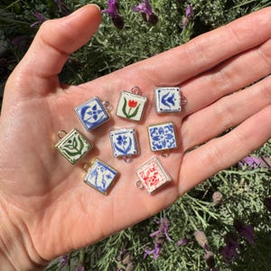 Op de afbeelding: Een verzameling vierkante keramische bedels met verschillende handgeschilderde ontwerpen, waaronder bloemen en geometrische patronen. Elke bedel heeft een zilver- of goudkleurig metalen frame en een kleine lus om aan sieraden of andere knutselwerken te bevestigen. De bedels worden in een hand gehouden.