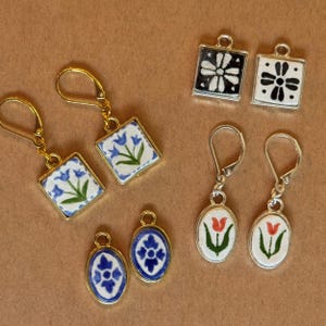 Può includere: Una collezione di orecchini fatti a mano con vari disegni. Alcuni orecchini presentano motivi floreali blu su sfondo bianco, mentre altri hanno disegni floreali in bianco e nero o tulipani rossi. Gli orecchini hanno dettagli in metallo dorato o argentato.