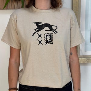 以下が含まれることがあります： ベージュの半袖Tシャツ。黒いグラフィックプリントが施されています。プリントには、走る犬、2つのスターバースト、切手のデザインが描かれています。シャツは柔らかく、ニュートラルカラーの生地で作られています。