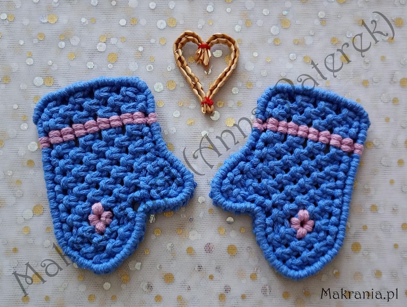May include: Two blue crochet mittens with pink stripes and a small pink flower detail. A woven heart is above the mittens. The background is a white fabric with gold and white dots. The text "Makrania.pl" is in the bottom right corner.