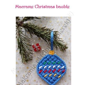以下が含まれることがあります： 赤、白、黄色のアクセントが付いた青いマクラメのクリスマスオーナメント。オーナメントは青いループから吊り下げられており、緑と白のステッチで装飾されています。画像には、小さな赤いギフトボックスと常緑樹の小枝も含まれています。画像には「Makrania.pl」と書かれています。