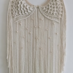 Op de afbeelding: Een witte macramé wandhanger met een houten ring en houten kralen. De macramé is in een franjes stijl met een geometrisch patroon.