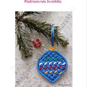 以下が含まれることがあります： 赤、白、黄色でカラフルなステッチが施された青いマクラメのクリスマスオーナメント。オーナメントは青いループから吊り下げられており、小さな赤いギフトボックスと松の枝と一緒に表示されています。「Makramowa bombka」というテキストが上部にあります。