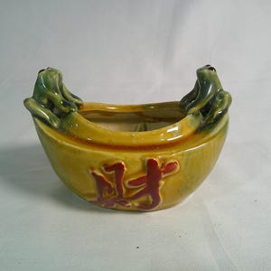 May include: A yellow ceramic bowl with a glossy finish, featuring two green frog figures perched on opposite sides of the rim. The front of the bowl displays raised Chinese characters in a dark red glaze, centered on the curved exterior surface.