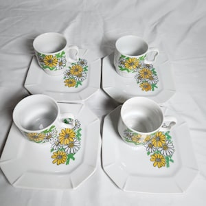 May include: Four white teacups and saucers. Each cup and saucer features a floral design with yellow and white daisies and green leaves. The saucers have a geometric, octagonal shape.