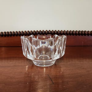 May include: A clear, faceted glass bowl with a scalloped rim. The bowl has a round base and a flower-like shape. The glass is transparent, allowing light to pass through. The bowl is sitting on a wooden surface.