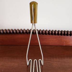 May include: A vintage metal potato masher with a green plastic handle. The masher has a three-pronged design and is in good condition.