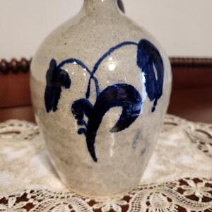 May include: A stoneware jug with a rounded body and a narrow neck. The jug is light gray with dark blue floral designs. The jug has a small handle and is sitting on a white lace doily.