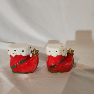 May include: A pair of red and white ceramic Christmas stocking salt and pepper shakers. The shakers have a white fur trim, green holly leaves, and a gold star on top.
