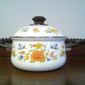 May include: White enamel cooking pot with a lid, featuring a floral design in orange, yellow, and blue. The pot has black handles and a dark brown knob on the lid. A vintage kitchenware item.