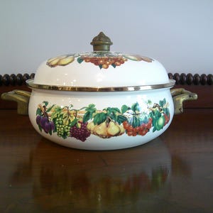 May include: A white enamelware pot with a lid, featuring a colorful fruit and vegetable design. The pot has gold-colored trim and handles. The lid is topped with a decorative knob. The pot is sitting on a dark wooden surface.
