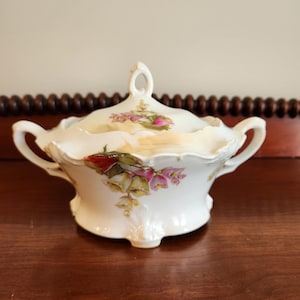 May include: White porcelain sugar bowl with lid, featuring a floral design with pink and red flowers. The bowl has two handles and a scalloped edge.