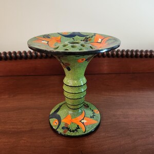 May include: A green ceramic flower frog vase with a speckled finish, featuring a wide, flat top with multiple circular holes for stems. The base and top are decorated with painted orange and black floral motifs and black horizontal line accents.