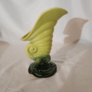 May include: A yellow ceramic seashell vase with a green base. The vase is shaped like a conch shell with a wide opening at the top. The base is decorated with waves.