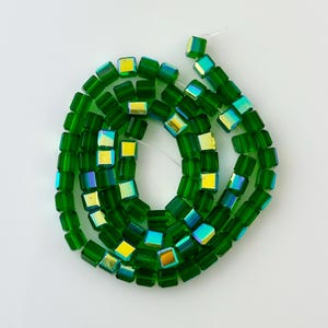 May include: A strand of green iridescent glass square beads. The beads are strung on a clear thread and are arranged in a circle.