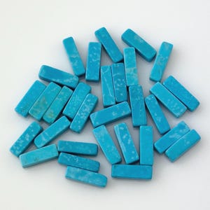 May include: A collection of rectangular turquoise-colored gemstone beads. The beads are a vibrant blue-green hue with white marbling. They are scattered on a white surface, showcasing their shape and color.