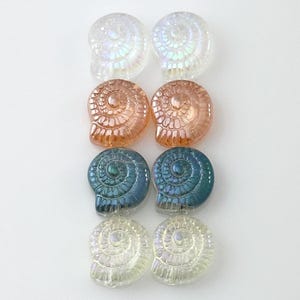 May include: An assortment of six iridescent seashell-shaped beads in various colors. The beads are arranged in three rows of two, with colors including clear, peach, blue-green, and pale yellow. The beads have a spiral design.