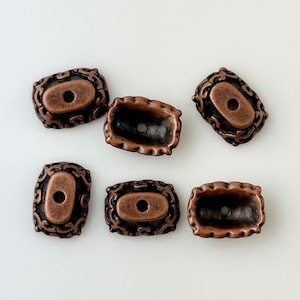 May include: Six antique copper-toned metal beads with ornate designs. Each bead has a hole in the center.