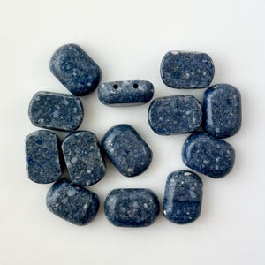 May include: A collection of twelve dark blue oval-shaped beads with white speckles. The beads have a smooth, polished finish.