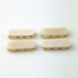 May include: Four rectangular, off-white beads with rounded edges and four evenly spaced holes. The beads are arranged in a square formation against a white background. These beads could be used for jewelry making or other craft projects.