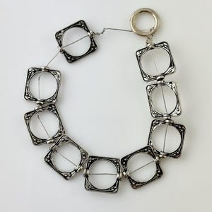 May include: A silver-toned bracelet featuring square, openwork links with decorative edges. The bracelet is strung on a thin wire and has a circular clasp. The design is symmetrical and the links are evenly spaced, creating a geometric pattern.