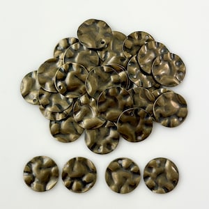 May include: A collection of bronze-colored, hammered metal disc charms. Each charm is round with a textured surface and a small hole for stringing. The charms are scattered on a white surface, suggesting their use in jewelry making or crafting.