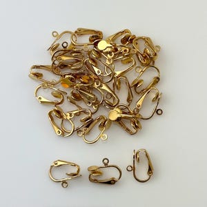 May include: A pile of gold-tone metal earring clips. The clips are designed to attach to the earlobe without piercing. There are three individual clips in the bottom right corner of the image.