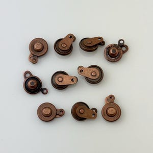 May include: Ten copper-colored metal slider buckles with a round button and a small loop on each side. The buckles are designed for attaching to clothing or other fabric items.