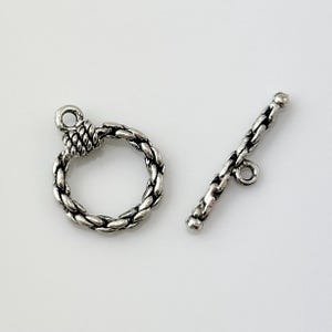 May include: Silver tone toggle clasp with a braided rope design. The clasp has a round loop and a straight bar with a small loop at one end.