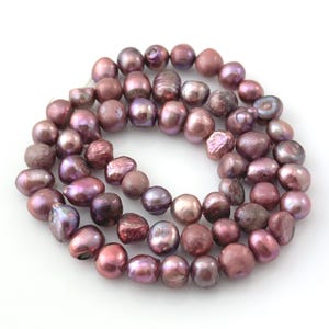 May include: A strand of freshwater pearls in shades of mauve, plum, and rose. The pearls vary in size and shape, with a mix of round and irregular forms. The pearls have a lustrous sheen, reflecting light and creating a subtle iridescence.