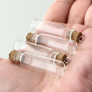May include: Four small, clear glass bottles with cork stoppers and silver-colored metal loops. The bottles are cylindrical and appear empty. The corks are a light brown color. The bottles are held in a person's hand.