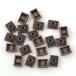 May include: A collection of small, rectangular copper-colored beads. Some beads feature a central circular design with surrounding ornate patterns, while others have a simple rectangular shape. The beads are scattered on a white surface.
