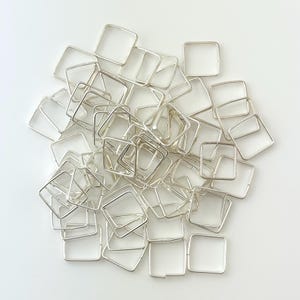 May include: A collection of small, square, silver-colored metal components scattered on a white surface. Each component is a hollow square, with clean lines and a bright, reflective finish. The arrangement is random, creating a visually interesting pattern.