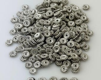 May include: A large pile of silver metal beads with a decorative, scalloped edge. The beads are arranged in a random pattern and are all the same size. There are also ten smaller beads lined up in a row at the bottom of the image.