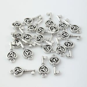 May include: A collection of small, silver-colored key charms. Each key features a decorative Celtic knot design on the head and a small key bit. The charms are scattered on a white surface.
