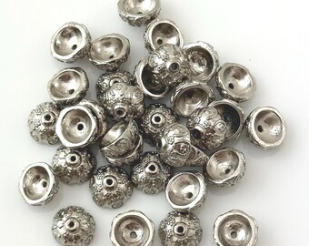 May include: A pile of silver metal beads with a decorative design. The beads are round and have a domed top.